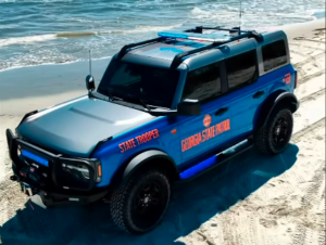 Blue and black SUV with "STATE LIFEGUARD" and "ORANGE COUNTY LIFEGUARD" text on beach sand near ocean waves