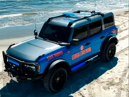 Blue and black SUV with "STATE LIFEGUARD" and "ORANGE COUNTY LIFEGUARD" text on beach sand near ocean waves