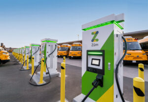 Row of green and white electric vehicle charging stations with yellow bollards; yellow taxis parked in background